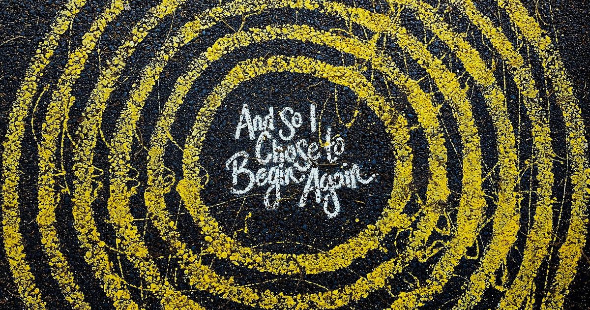 yellow paint circles on black tarmac surrounding the words 'and so I choose to begin again'