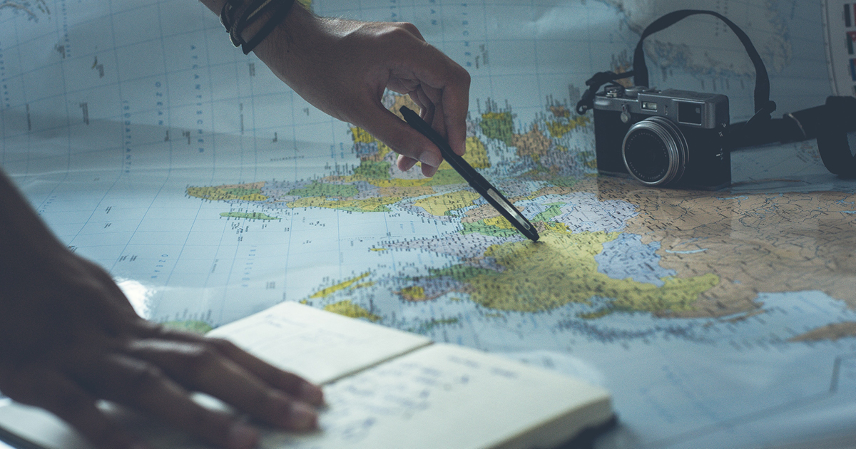 image shows a map laid out with a camera placed on top and a persons hand with a pen pointing to an area on the map.